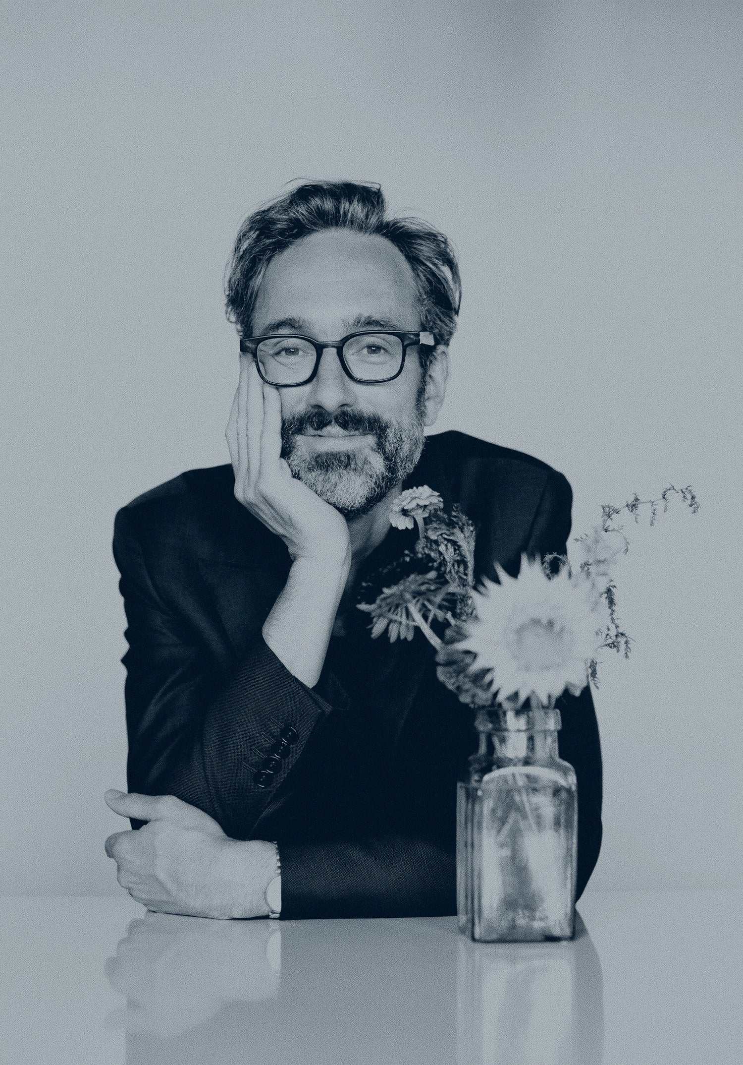 Black and white image of roots artist David Myles leaning on his hand at a white shiny table with wildflowers in a vase next to him