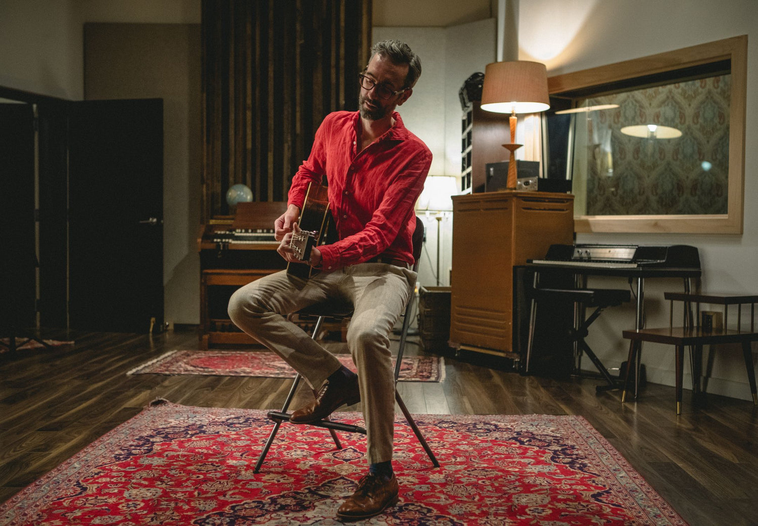 David Myles sitting on a tall chair in the middle of an ornate room with guitar in lap playing in a red shirt and beige trousers. 