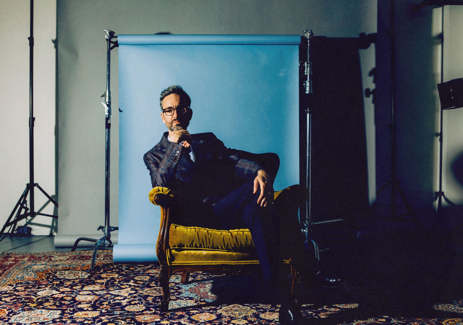 David Myles in a Nova Scotia studio in a yellow velvet chair leaning to one side in front of a blue screen and ornate rug on the floor.