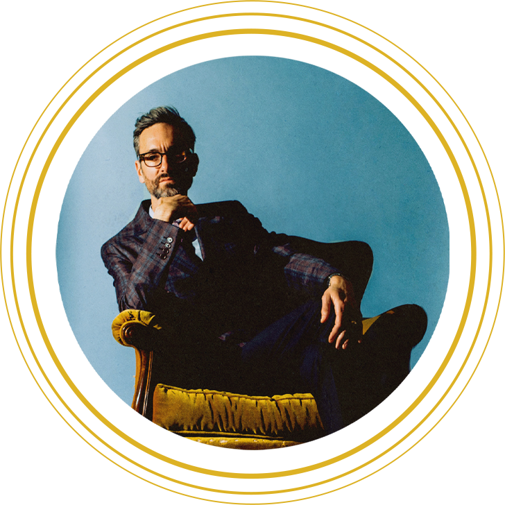 Circular image of musician David Myles leaning to one side in yellow armchair with a blue background wearing a checked suit and leaning on one arm.